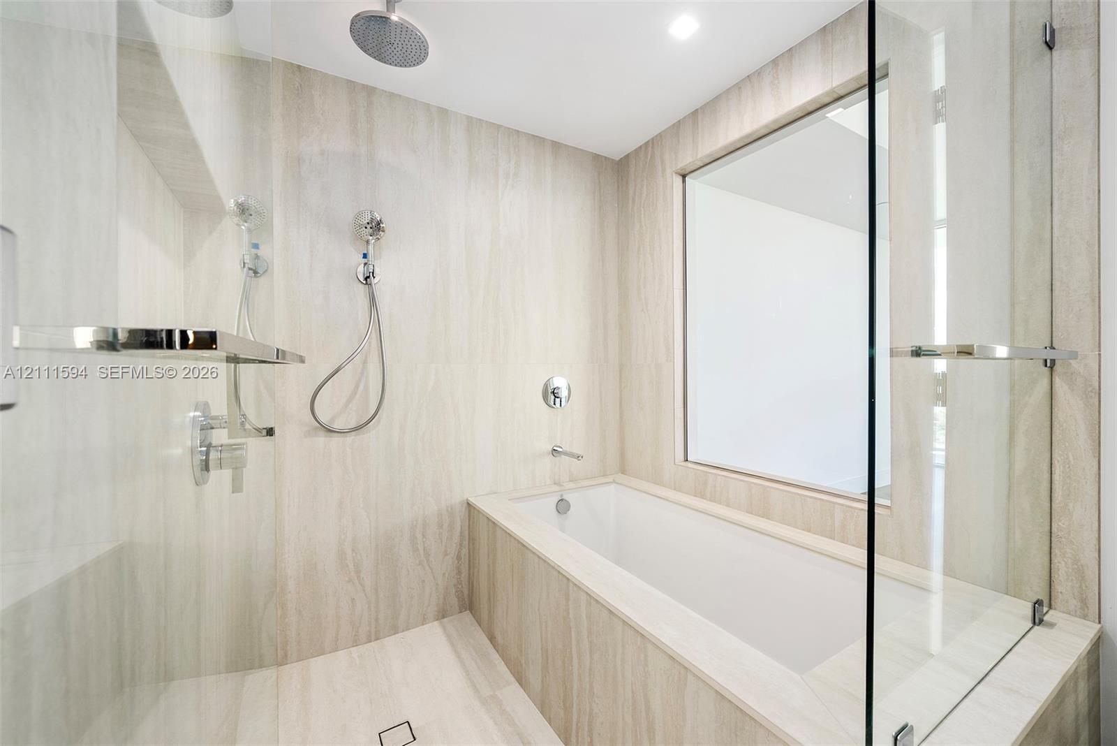 Wet room featuring stone-finish wall and floor tiling, deep-soaking bathtub, glass partition, wall-mounted shower system with rain head, and integrated shelving