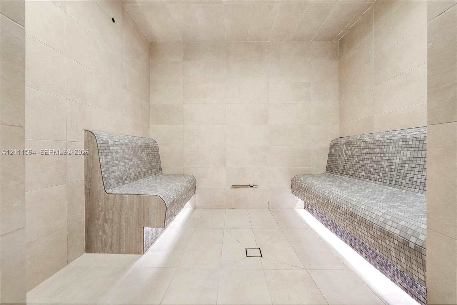 Steam room featuring large-format stone wall and floor tile, built-in dual benches with mosaic tile surfacing, integrated under-bench lighting, and a centered floor drain