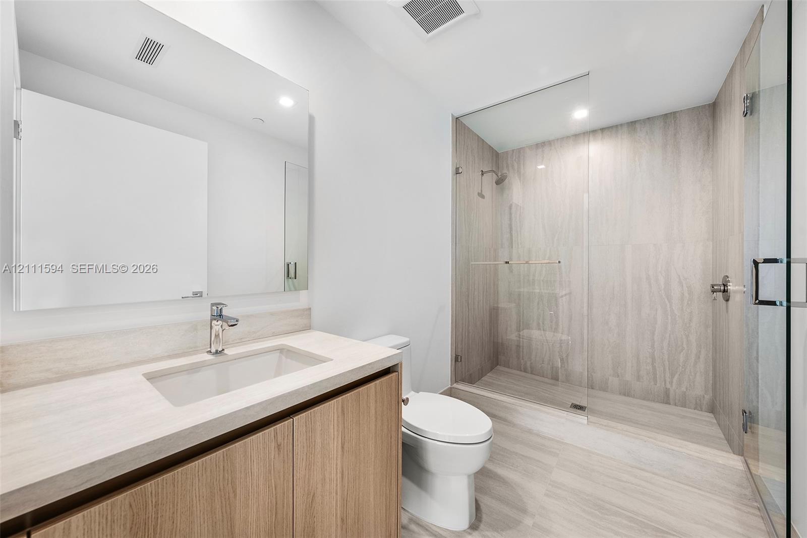 Bathroom featuring a wood-finish vanity with an integrated countertop, a frameless glass-enclosed walk-in shower with floor-to-ceiling stone-style tile, and a wall-mounted mirror