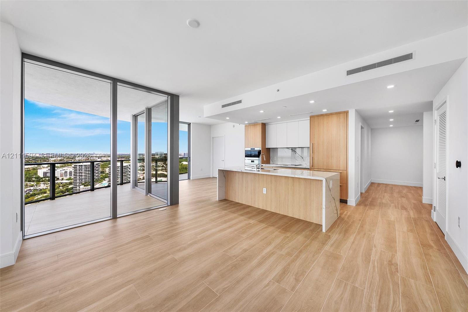 Open-concept interior featuring wood-finish flooring, floor-to-ceiling glass panel walls, a wraparound balcony, a kitchen island with stone-finish countertops, and minimalist flat-panel cabinetry