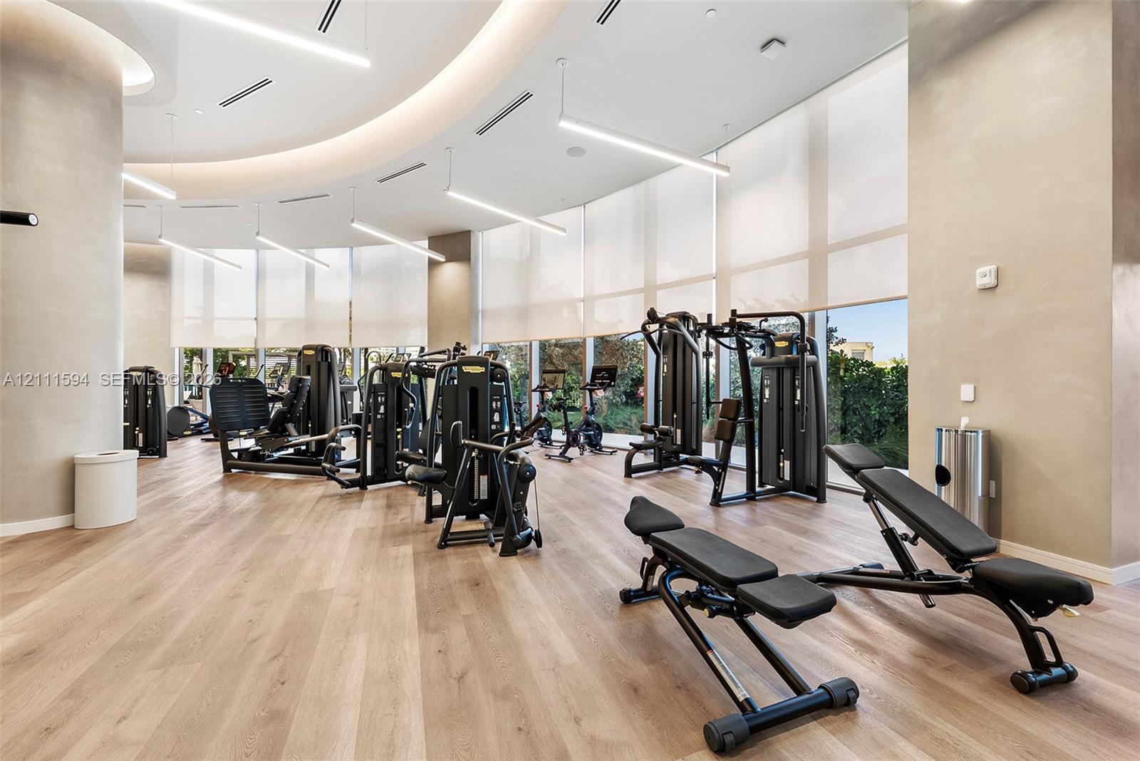 Fitness center featuring wood-finish flooring, floor-to-ceiling windows with roller shades, recessed cove lighting, and suspended linear LED fixtures