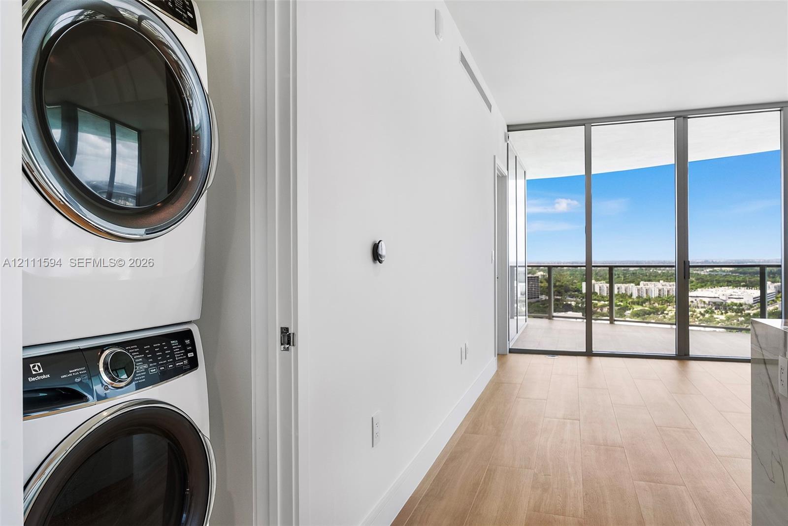 Stacked laundry appliances, wood-finish flooring, floor-to-ceiling windows, and private balcony