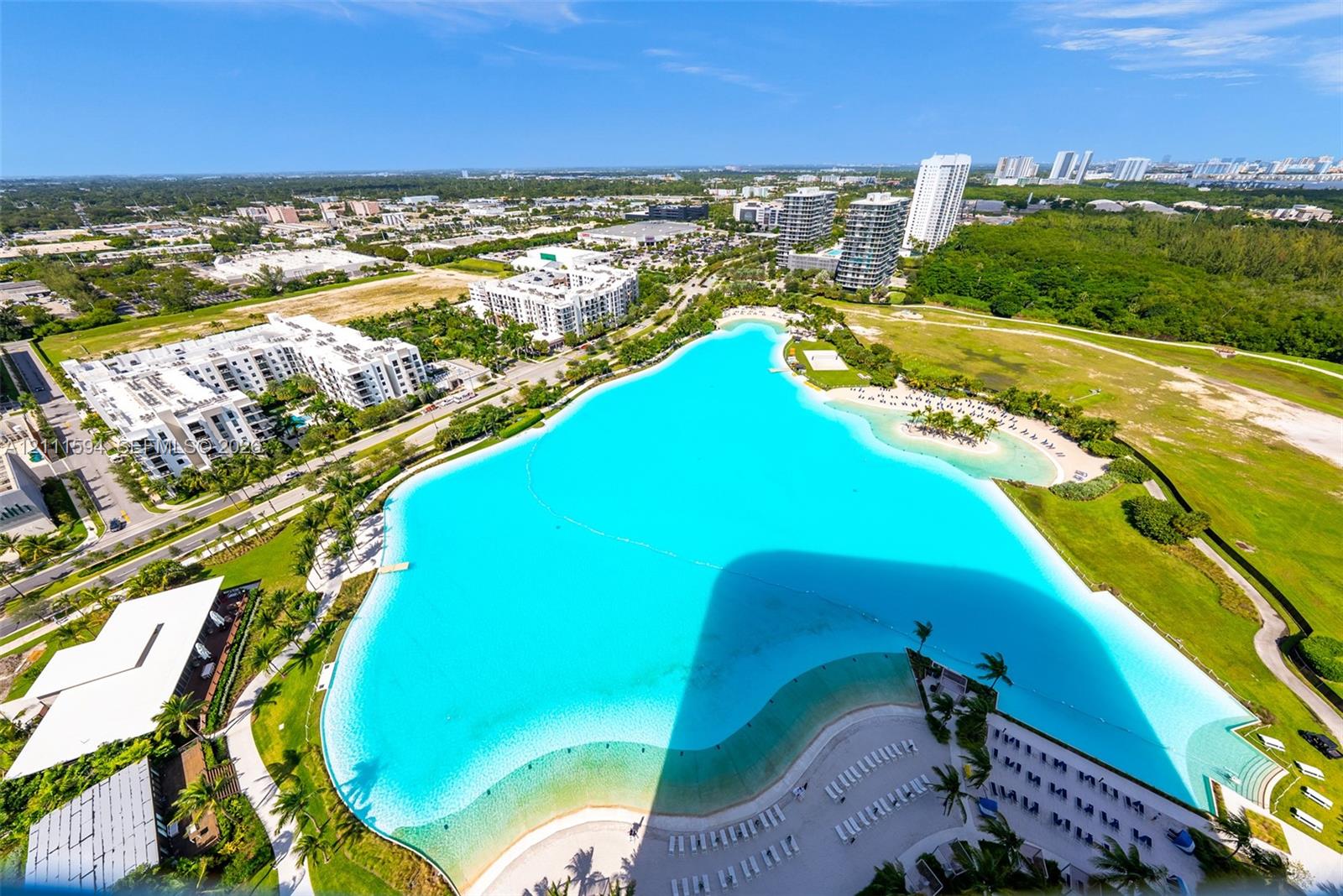 Aerial perspective featuring a large crystalline lagoon, expansive sandy shoreline, manicured green space, and paved pathways