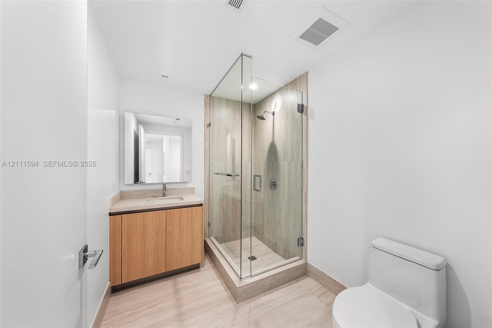 Walk-in shower with frameless glass enclosure and tile surround, wood-finish cabinetry, rectangular under-mount sink, and neutral tile flooring