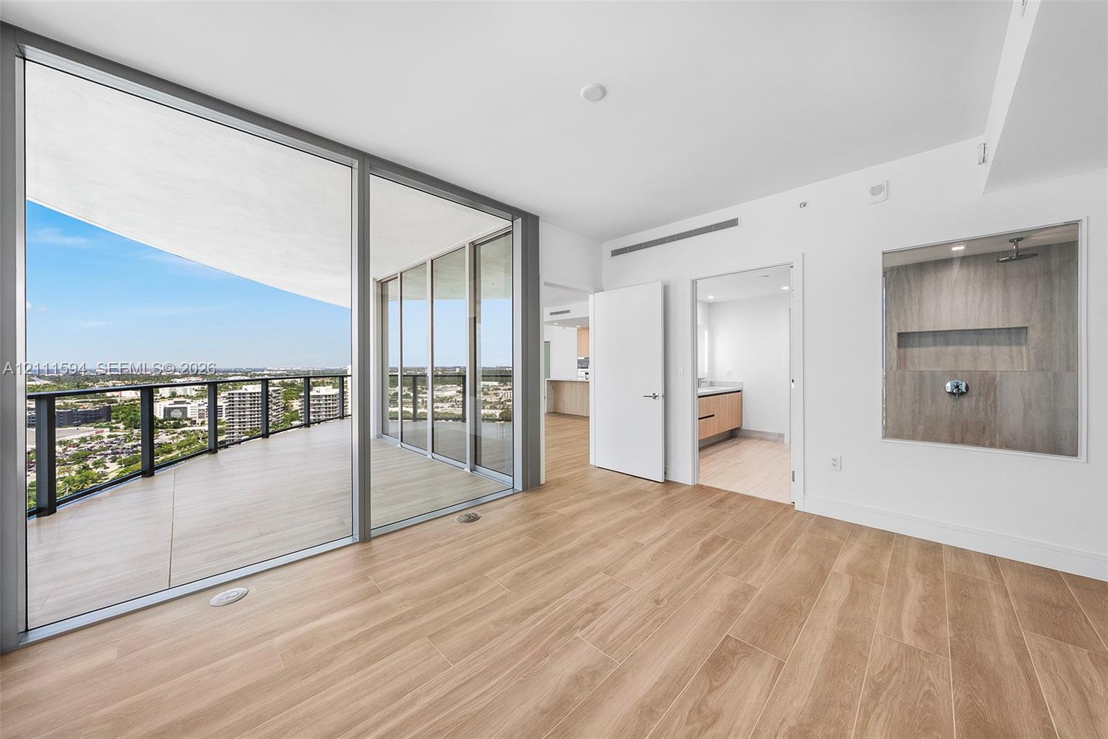 Floor-to-ceiling glass panel doors, expansive covered balcony, wood-finish flooring, and built-in glass-enclosed shower featuring stone-finish wall tile and recessed shelving