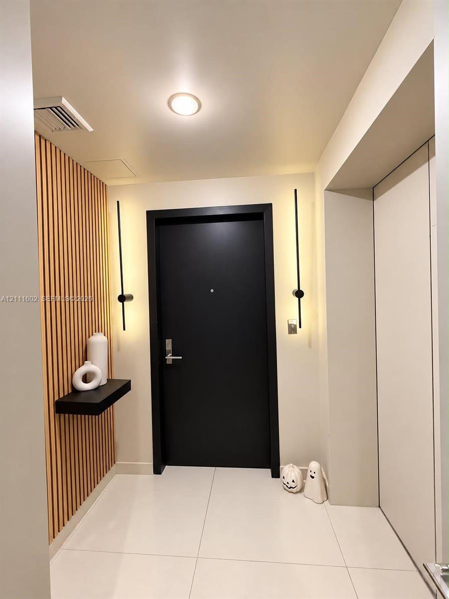 Entryway featuring vertical wood slat paneling, large-format tile flooring, a matte black door, dual linear wall-mounted light fixtures, and a floating shelf