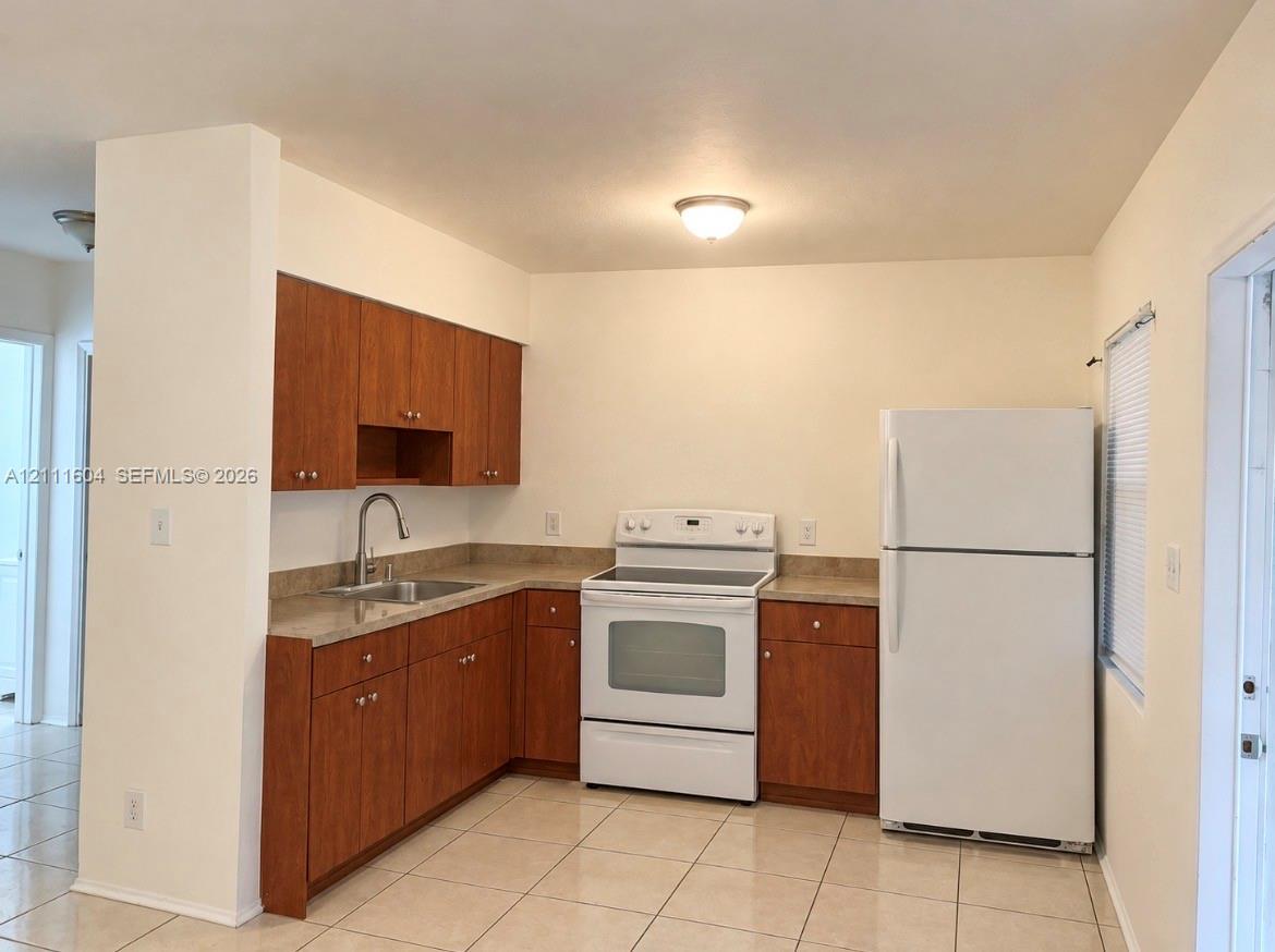 Kitchen featuring wood-finish cabinetry, neutral solid-surface countertops, tile flooring, and white kitchen appliances