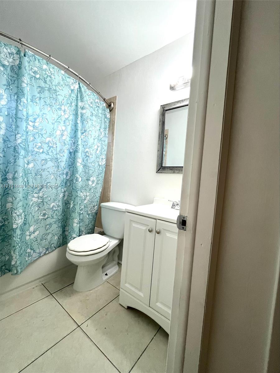 Bathroom featuring white vanity cabinetry, light-toned tile flooring, curved shower rod, and wall-mounted lighting fixture