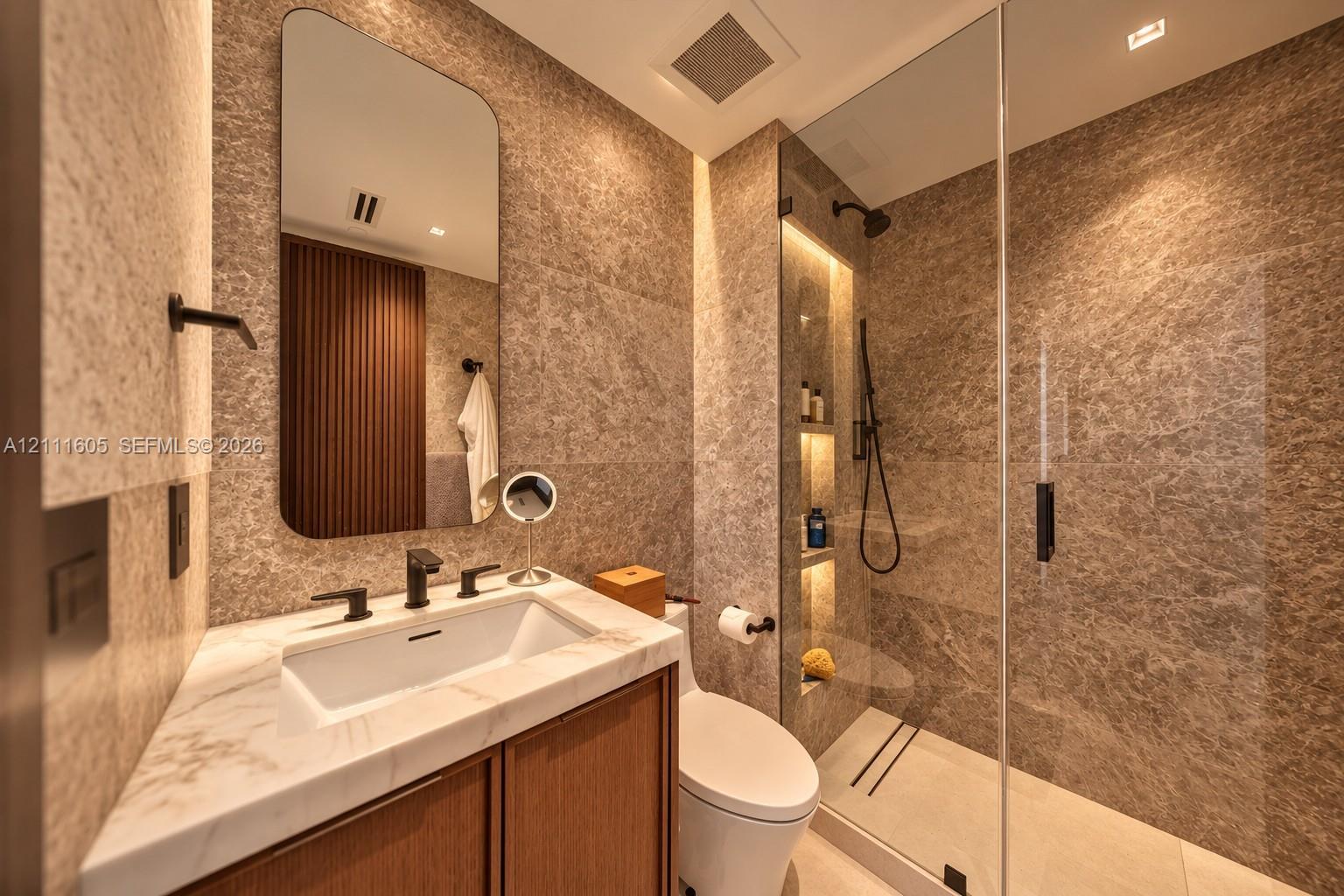 Bathroom featuring floor-to-ceiling stone tile, glass-enclosed shower with integrated recessed shelving, wood-finish vanity cabinet, stone countertop with undermount sink, and matte black fixtures