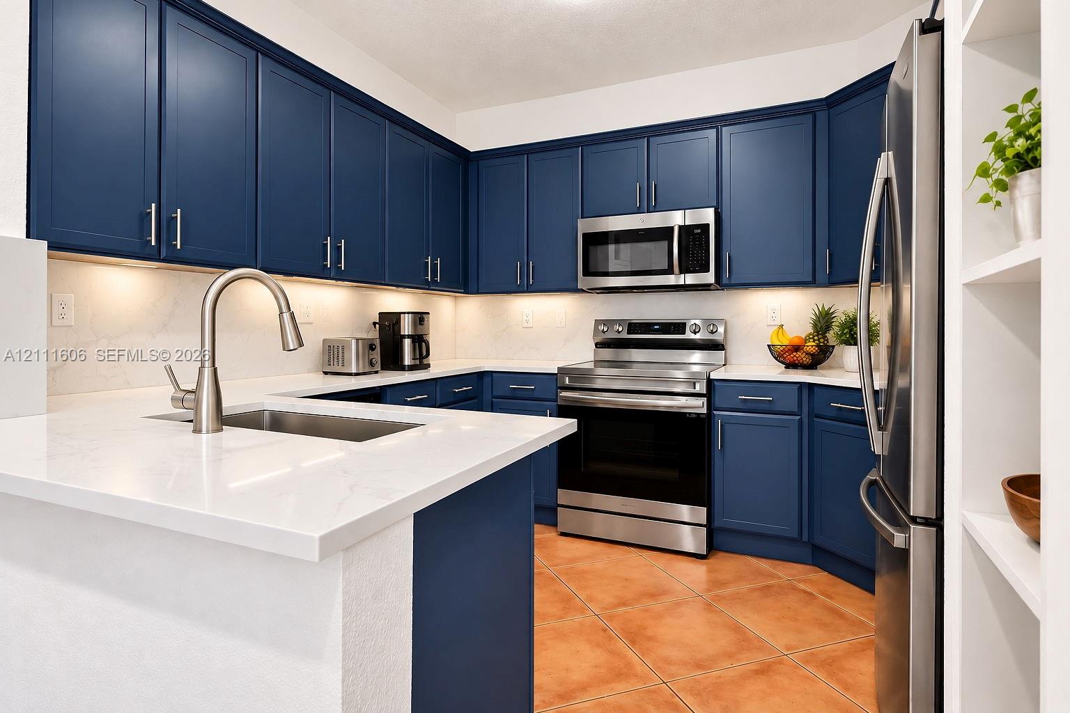 Kitchen featuring navy blue cabinetry, stainless steel appliances, light-toned stone-finish countertops, under-cabinet lighting, and ceramic tile flooring Kitchen featuring navy blue cabinetry, stainless steel appliances, light-toned stone-finish countertops, under-cabinet lighting, and ceramic tile flooring