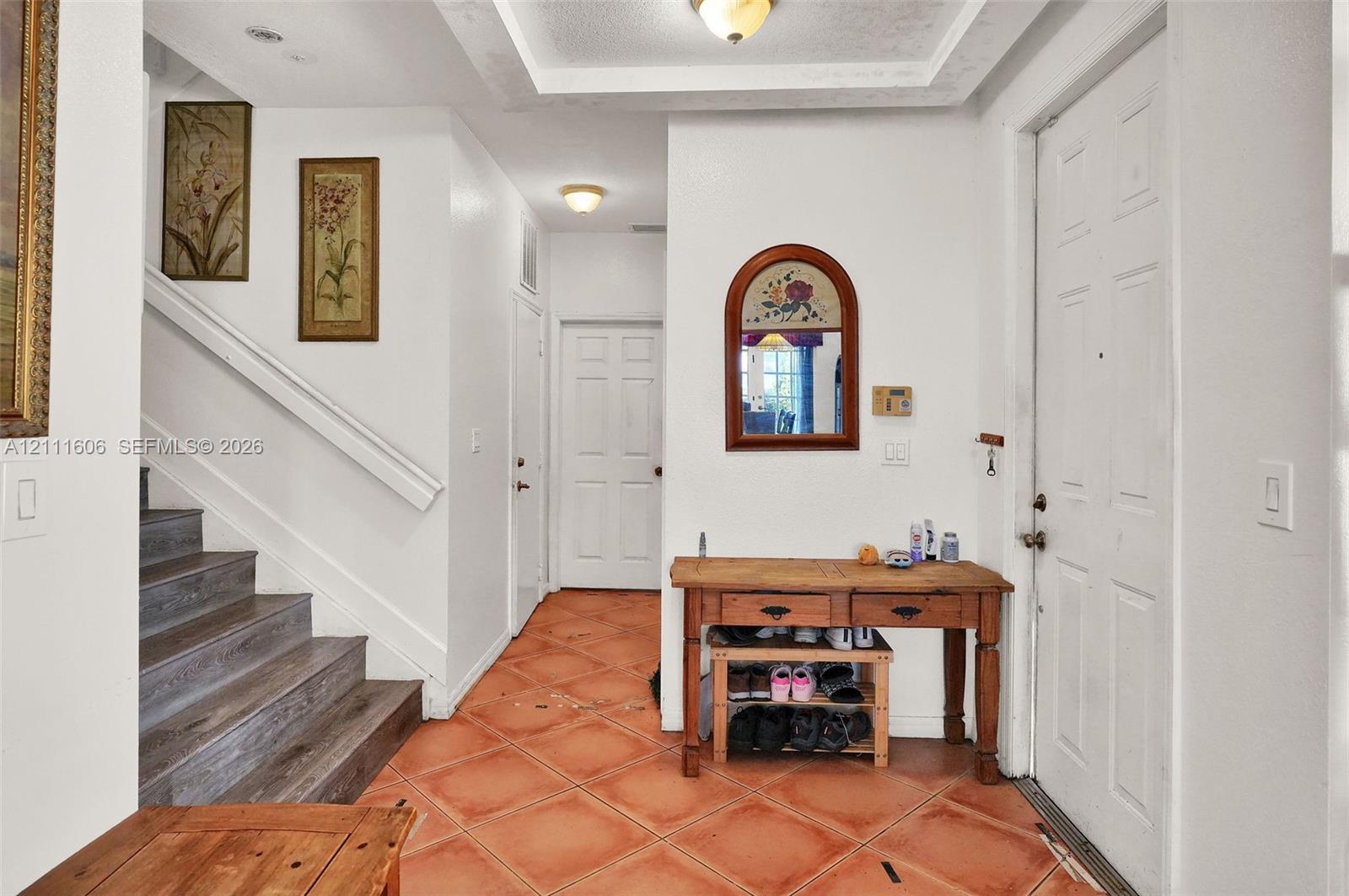 Foyer featuring terracotta tile flooring, recessed tray ceiling, built-in staircase with wood-finish treads, and arched wall-mounted mirror Foyer featuring terracotta tile flooring, recessed tray ceiling, built-in staircase with wood-finish treads, and arched wall-mounted mirror