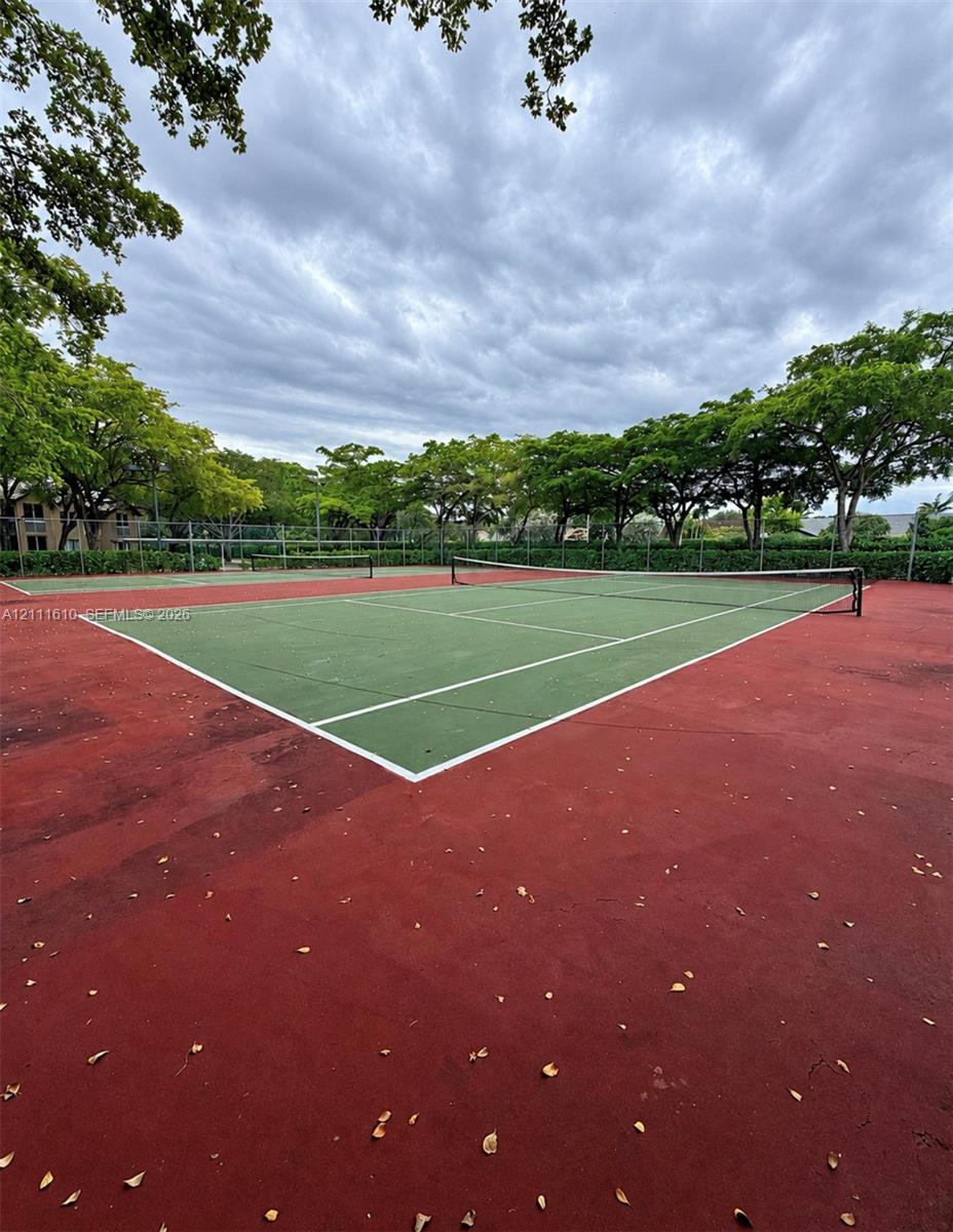 Dual tennis courts featuring a synthetic hard-court surface, perimeter chain-link fencing, and mature trees