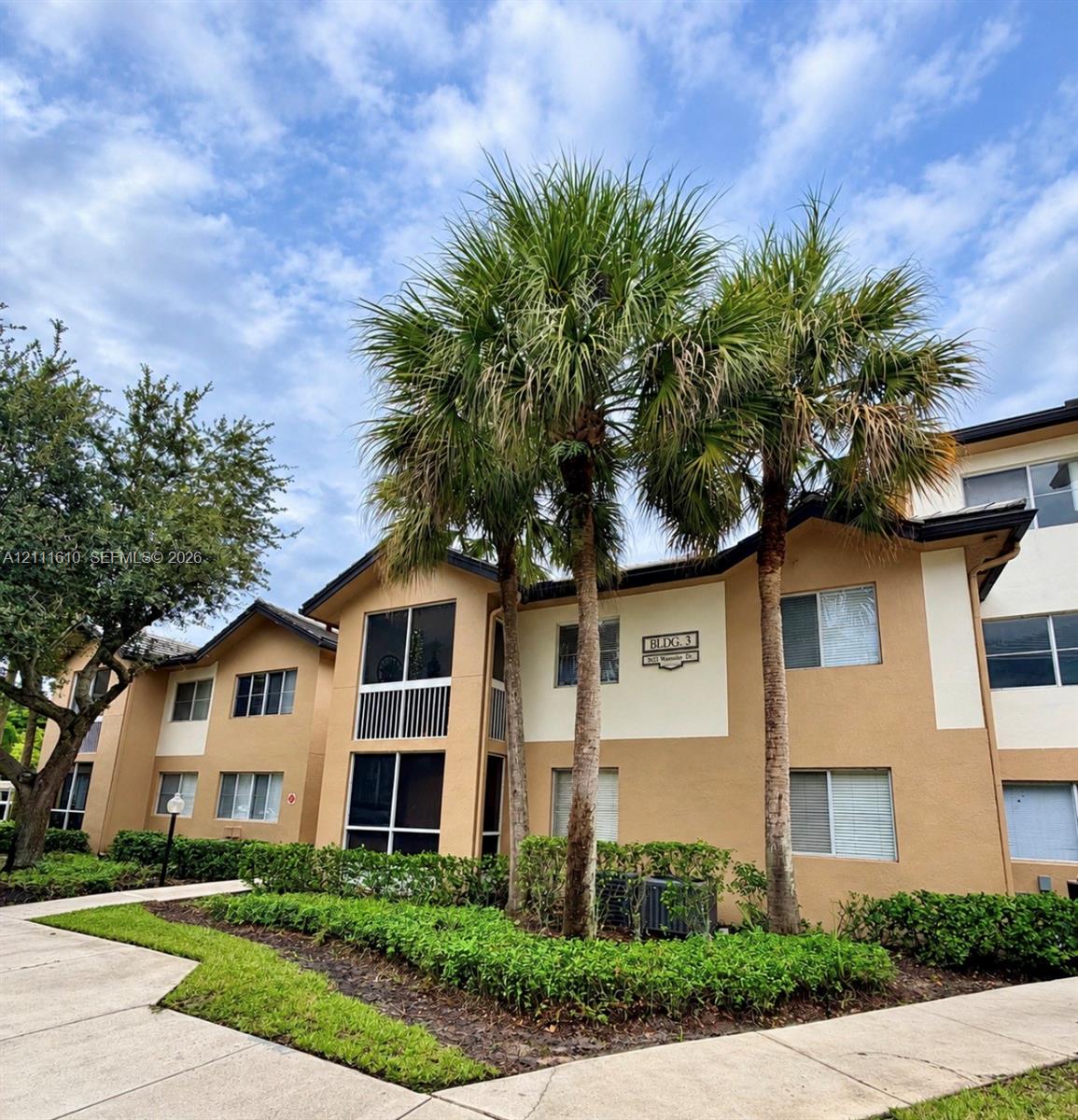 Multi-story exterior featuring a neutral stucco facade, screened-in balconies, mature palm trees, manicured hedging, and concrete walkways