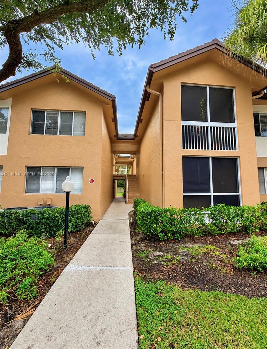 Stucco exterior featuring dual buildings, a concrete walkway, screened-in balconies, and manicured shrubbery