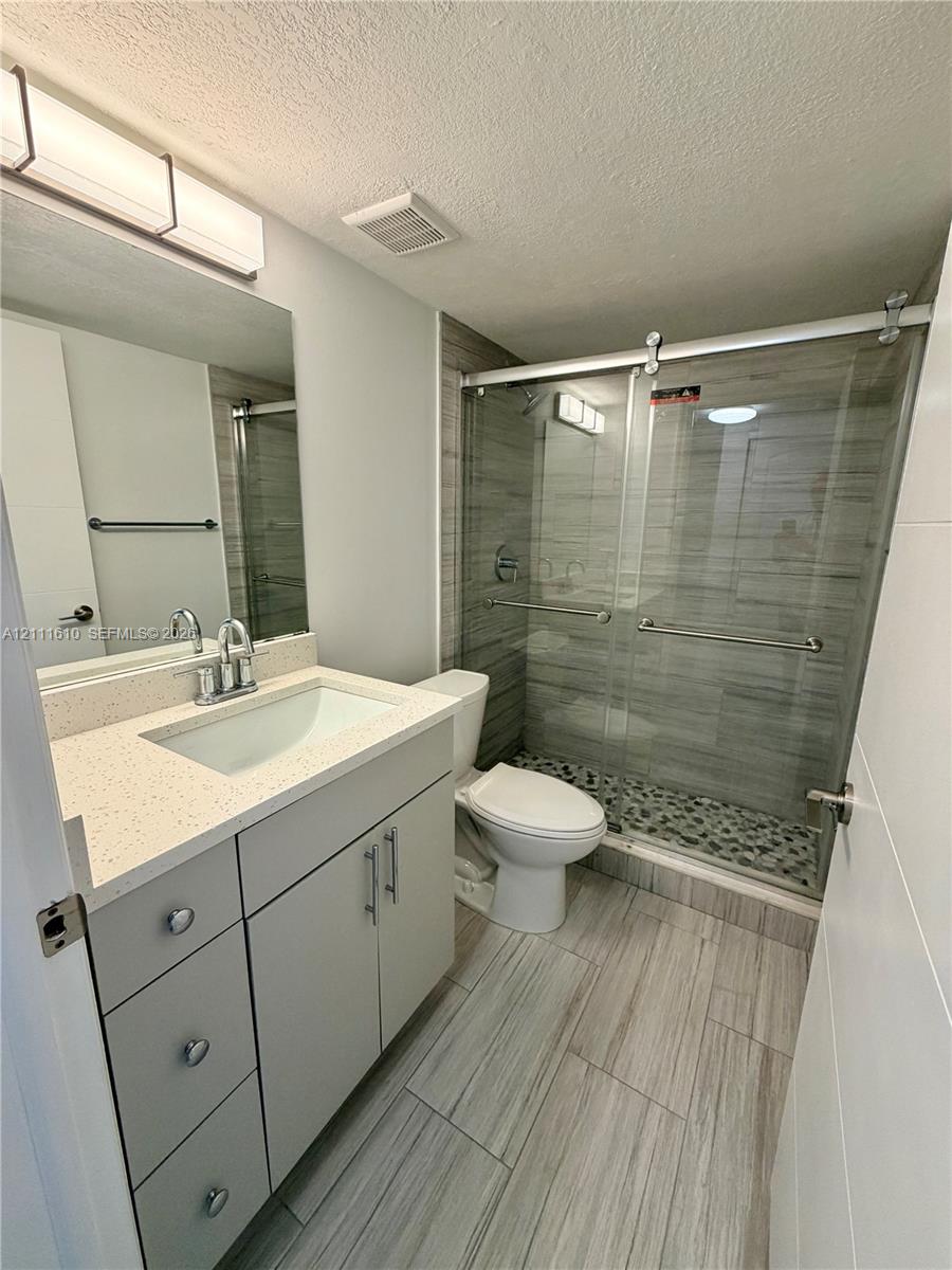 Bathroom featuring a quartz-topped vanity with an undermount sink, walk-in shower with a sliding glass door and pebble-stone floor, wood-finish floor tile, and wall-mounted vanity lighting