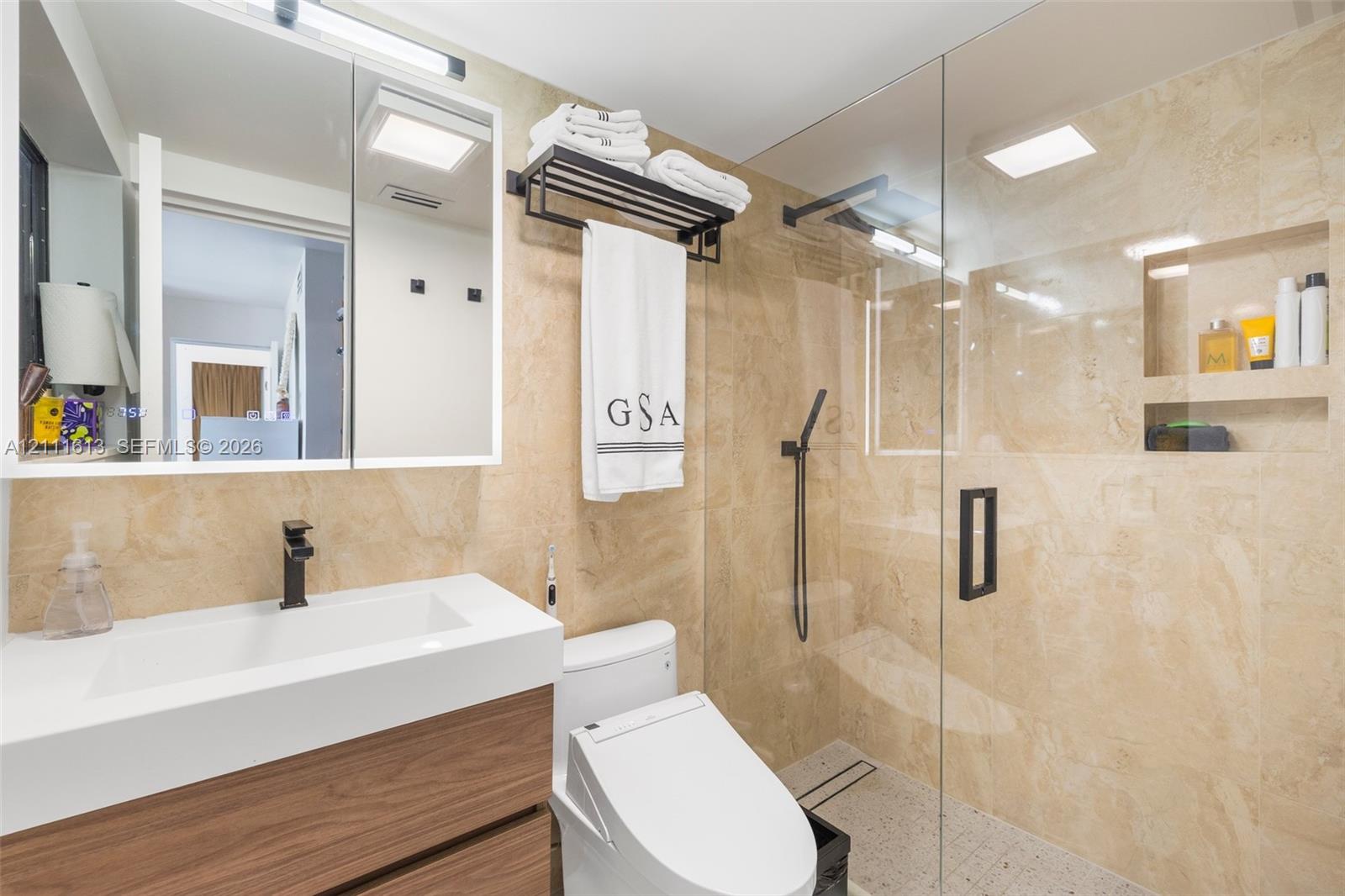 Bathroom featuring a wood-finish floating vanity with an integrated white sink, stone-tiled wall surfaces, a glass-enclosed shower with an integrated recessed shelf, and matte black hardware