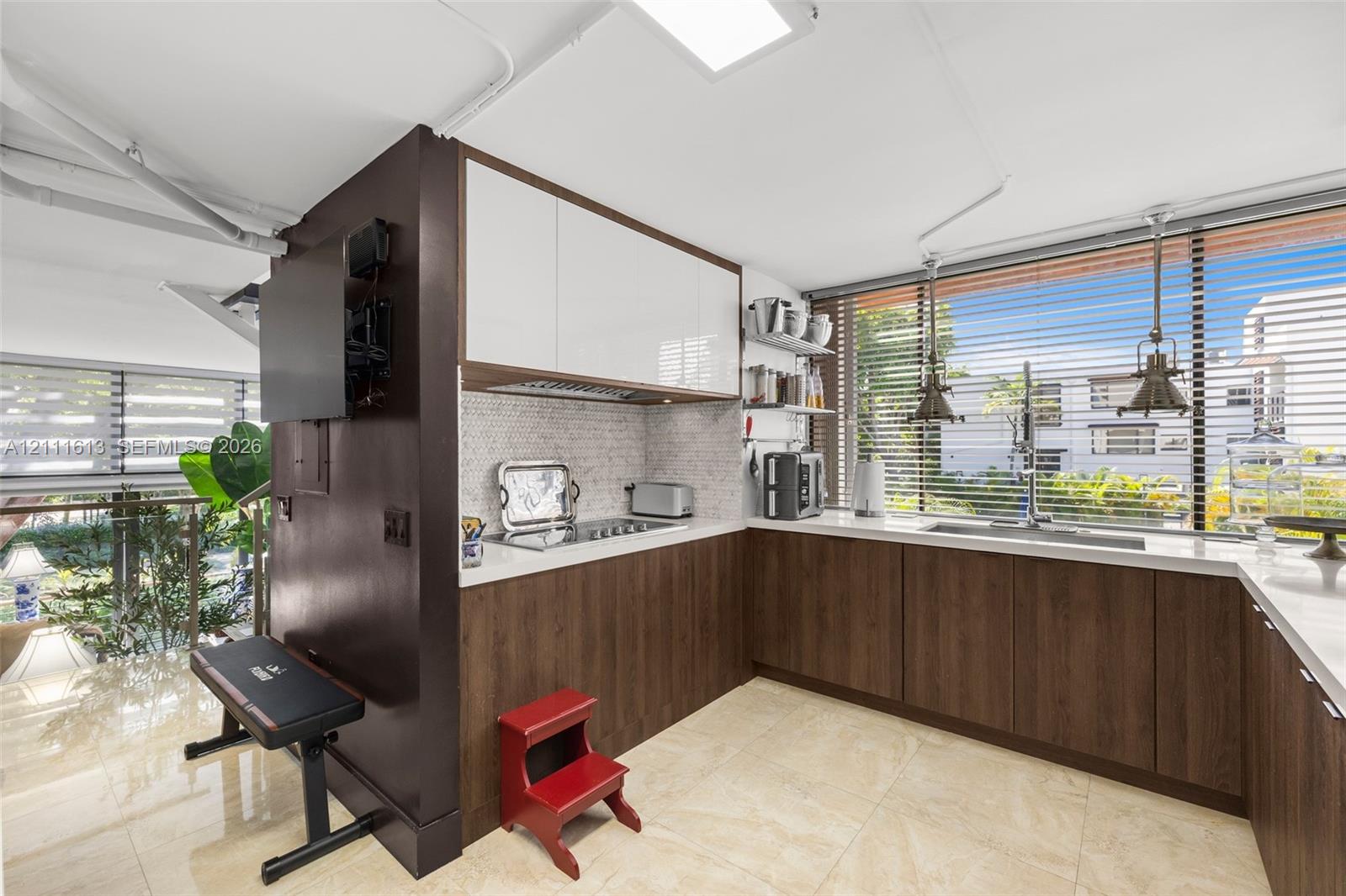 Kitchen featuring two-tone cabinetry with wood-finish lower units and high-gloss upper cabinets, solid-surface countertops, mosaic tile backsplash, and oversized windows with pendant lighting