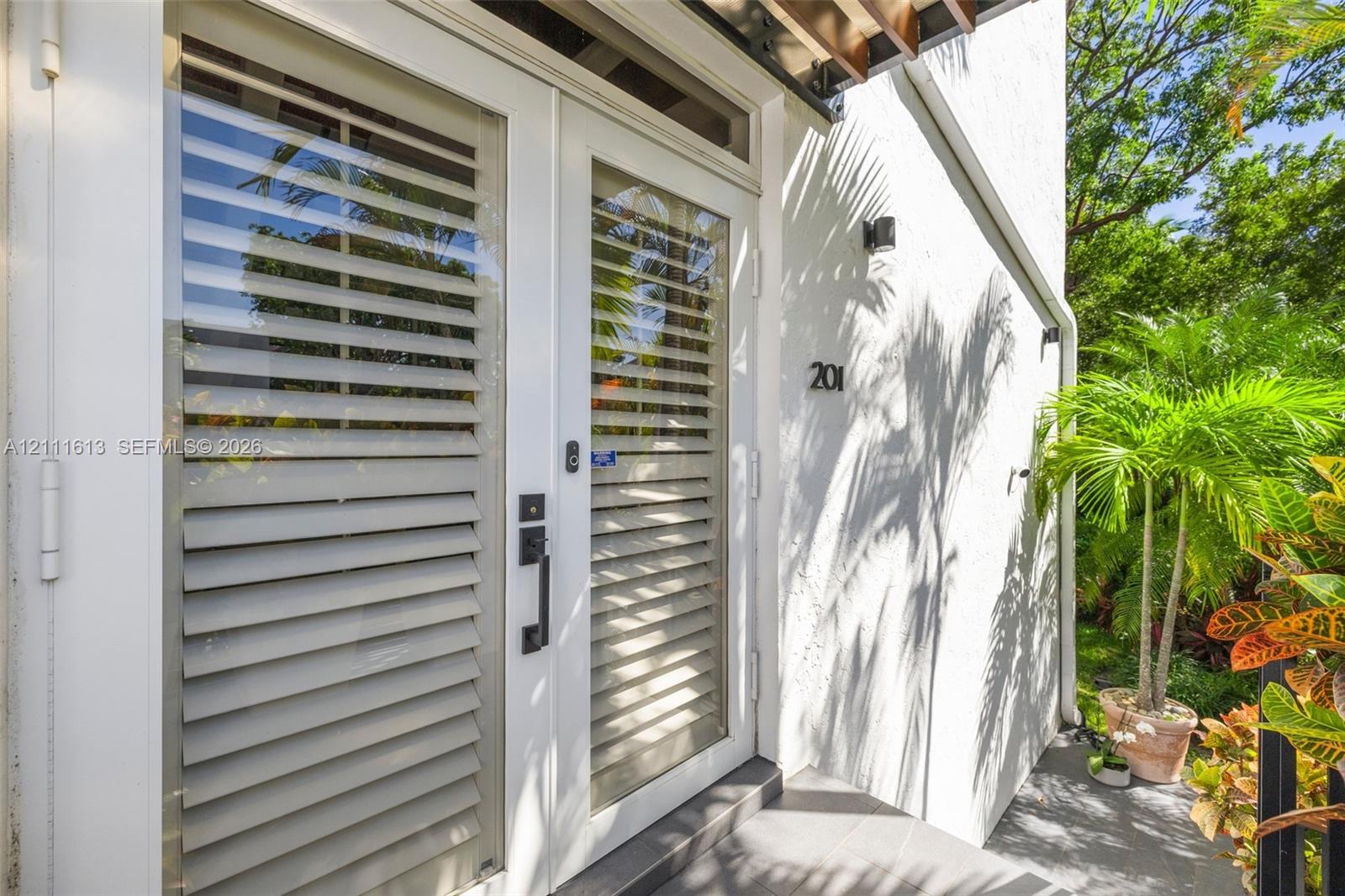 Exterior entrance featuring double glass-paneled doors with integrated horizontal louvers, a transom window, matte black hardware, and an overhead wooden pergola element Exterior entrance featuring double glass-paneled doors with integrated horizontal louvers, a transom window, matte black hardware, and an overhead wooden pergola element
