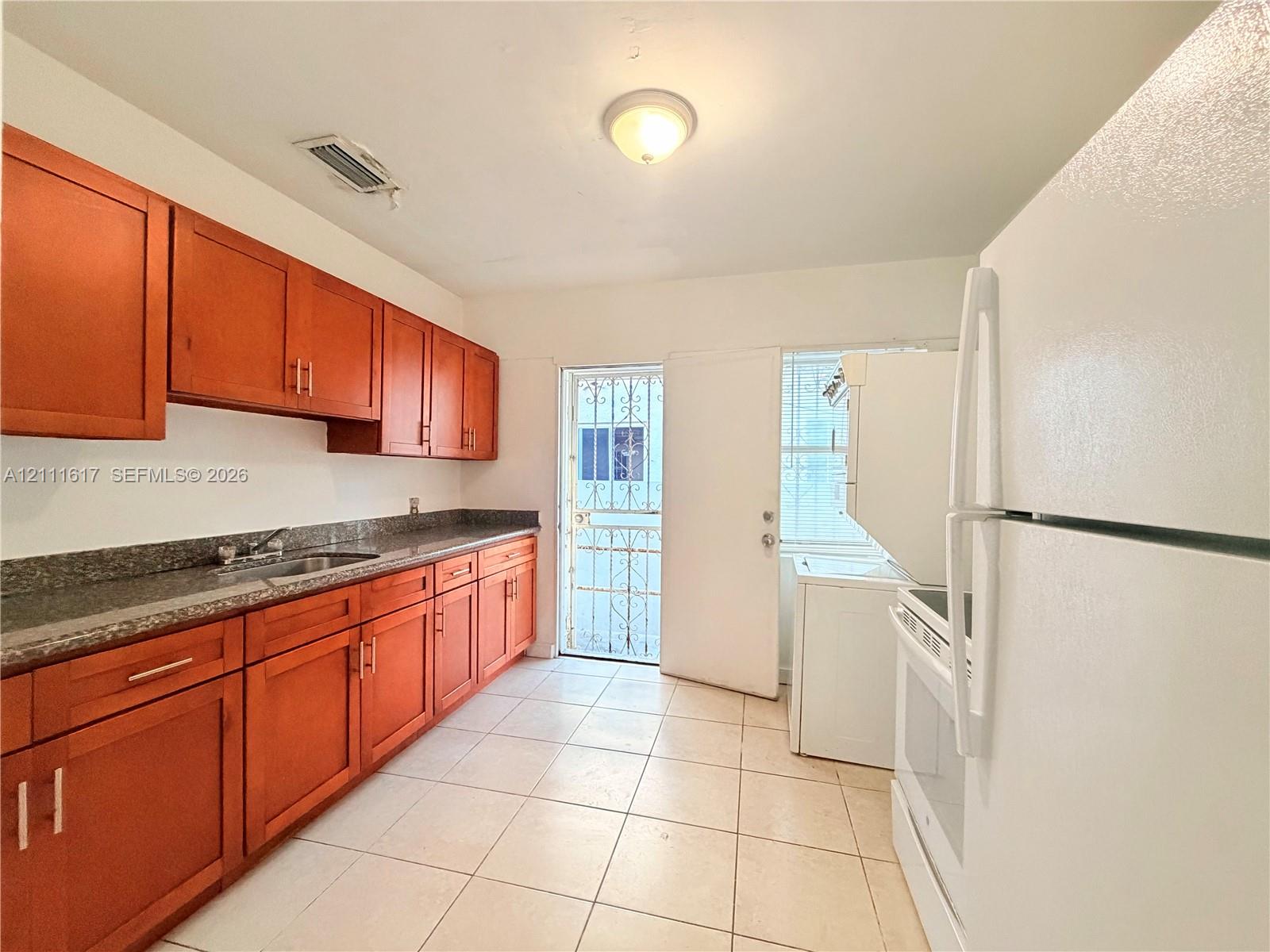 Kitchen featuring wood cabinetry, stone-finish countertops, stainless steel sink, tiled flooring, and exterior access via a metal security door