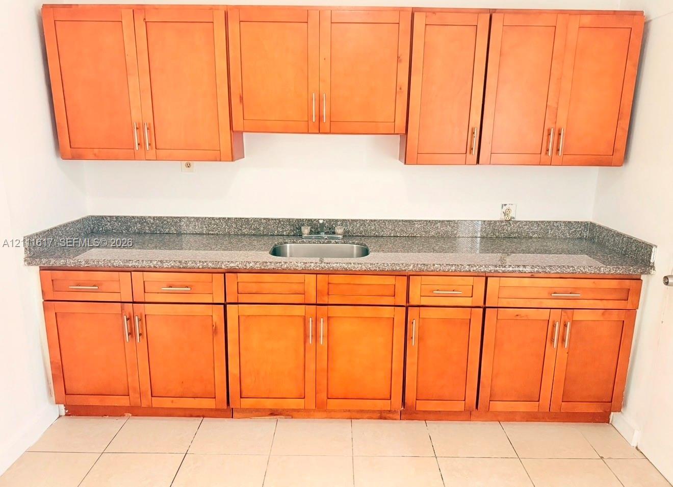Natural wood-finish cabinetry featuring shaker-style doors and metal hardware, stone-composite countertop, stainless steel undermount sink, and tile flooring
