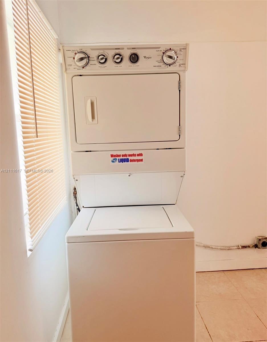 Stacked white washer and dryer unit with top-loading washing machine, ceramic tile flooring, and horizontal window blinds