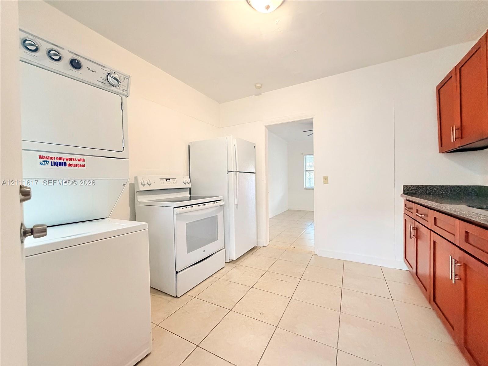 Kitchen featuring tile flooring, wood-finish cabinetry, dark solid-surface countertops, and a stacked laundry unit