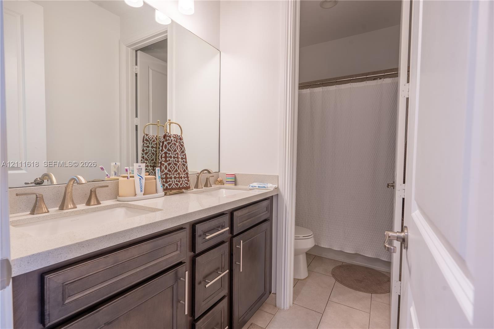 Bathroom featuring a dual-sink vanity with light-toned solid surface countertops, dark-finish cabinetry, metallic hardware, and a tub-shower combination with tile flooring