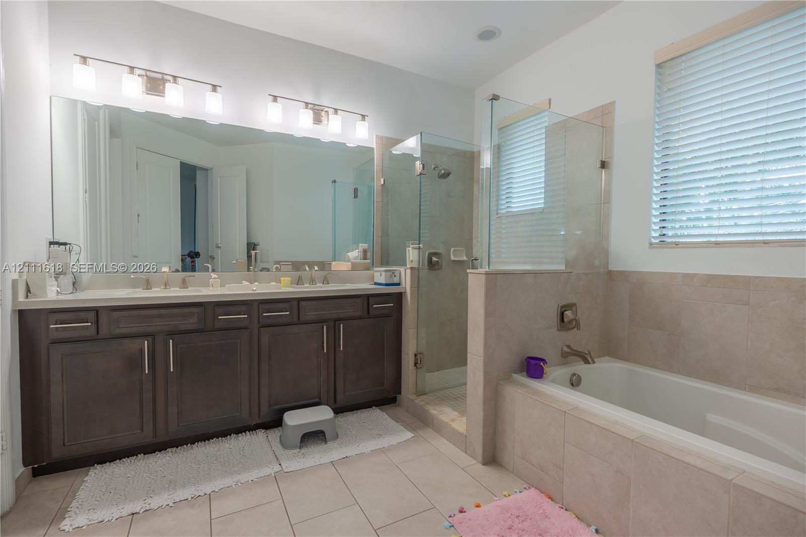 Double vanity with dark cabinetry, large wall-to-wall mirror, glass-enclosed shower, and deep-soaking bathtub with tile surround