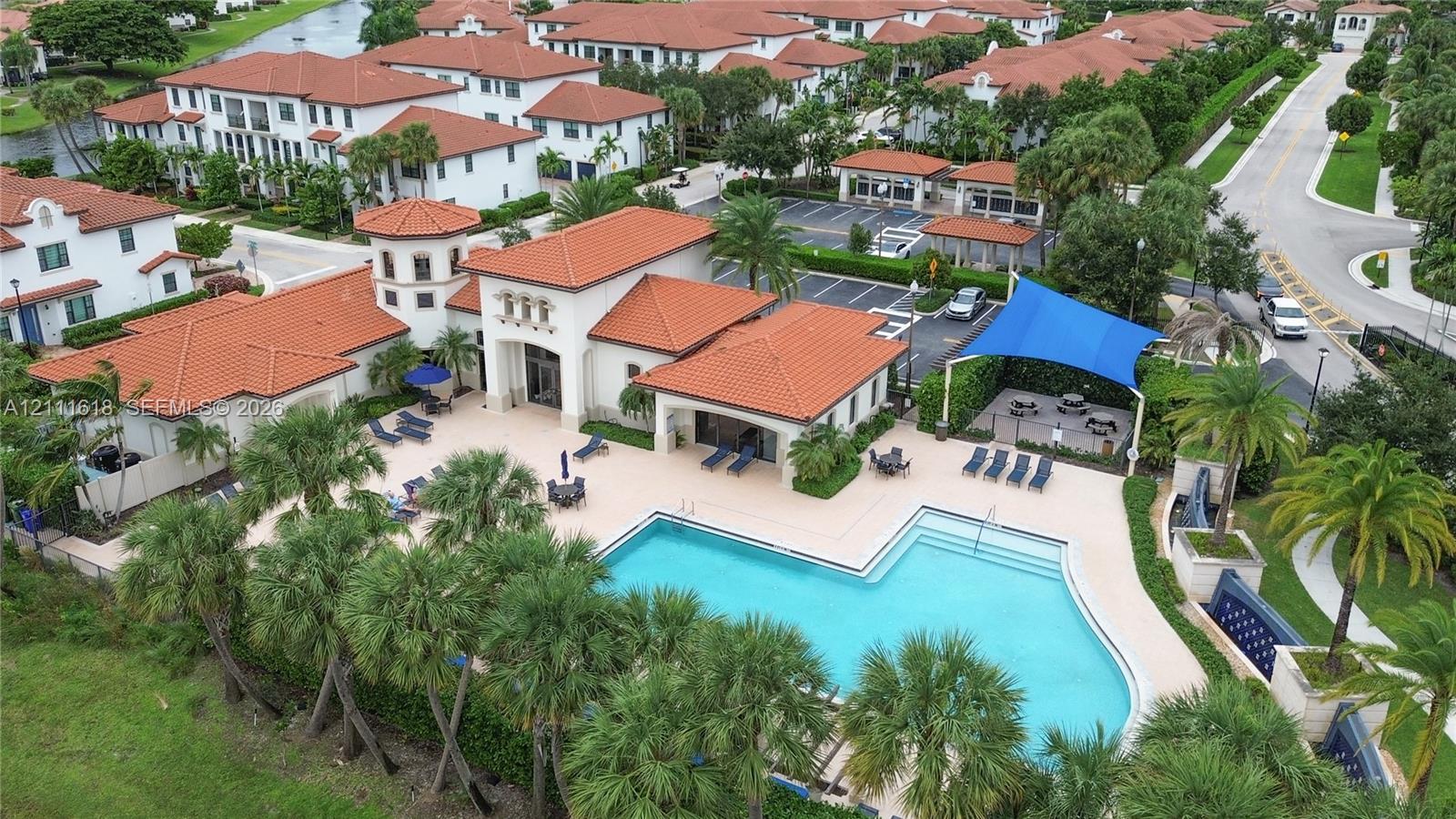 Community center featuring a swimming pool, clay-tile roof architecture, expansive pool deck, shaded canopy area, and mature palm landscaping