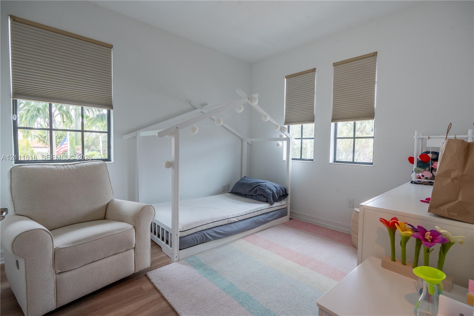 Bright bedroom featuring wood-finish flooring, three windows with grid-pattern muntins, and neutral cellular shades
