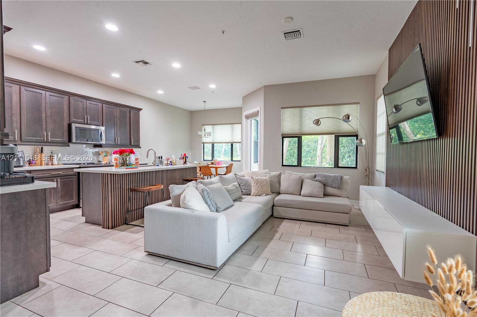 Open-concept floor plan featuring dark wood-finish cabinetry, a kitchen island with vertical wood detailing, large-format tile flooring, and a floor-to-ceiling slatted wood accent wall
