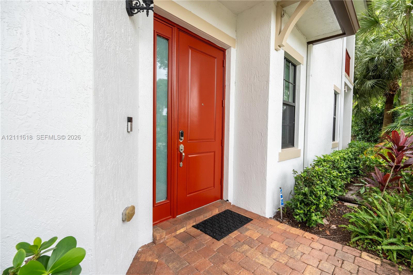 Exterior entryway featuring a red solid-core front door with an integrated sidelight, stucco facade, brick paver flooring, and decorative corbel accents