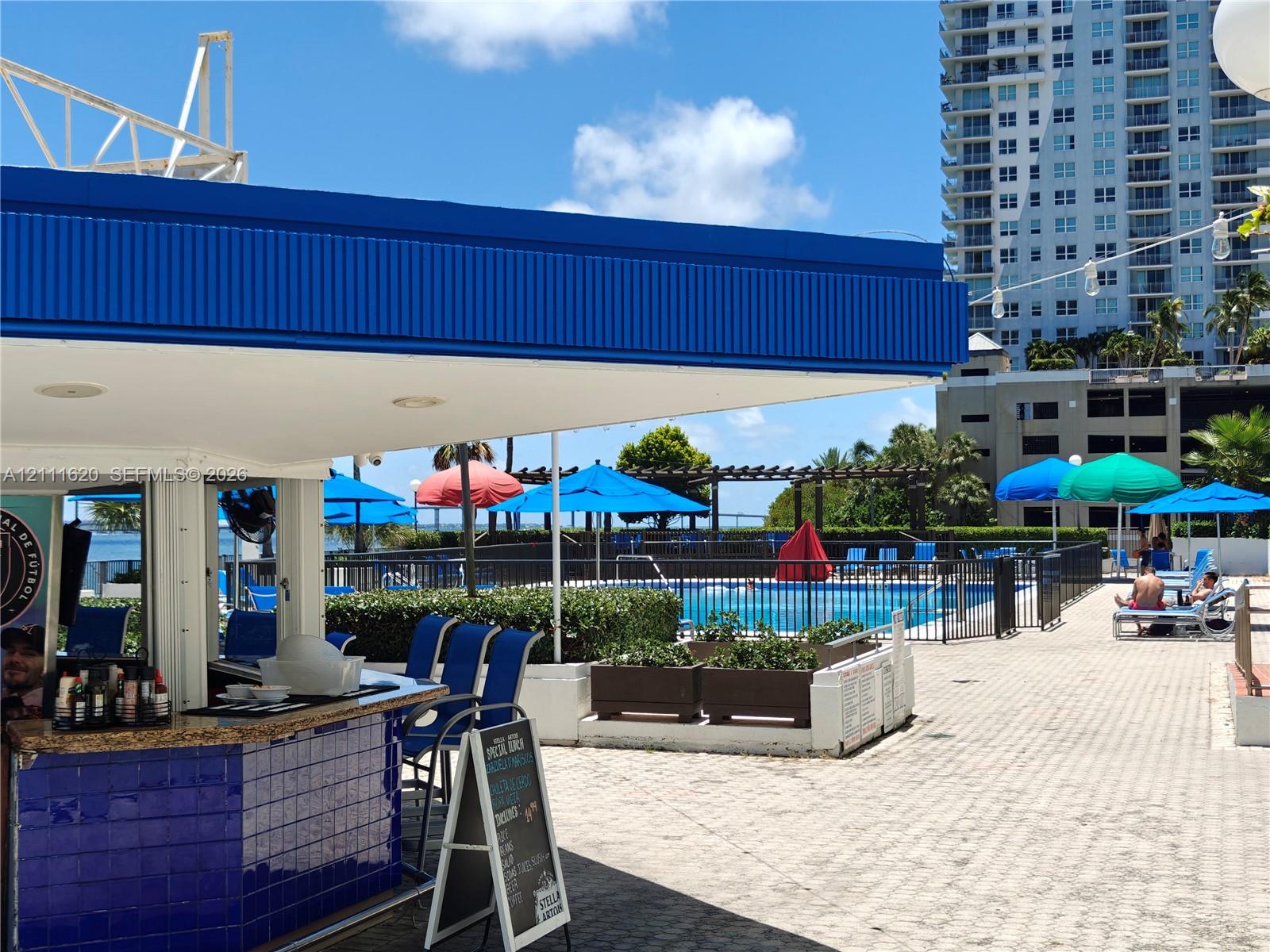 Outdoor bar featuring blue tile facing, stone-finish countertop, and recessed ceiling lighting, situated on a paver deck with an integrated swimming pool, metal perimeter fencing, and raised concrete planters