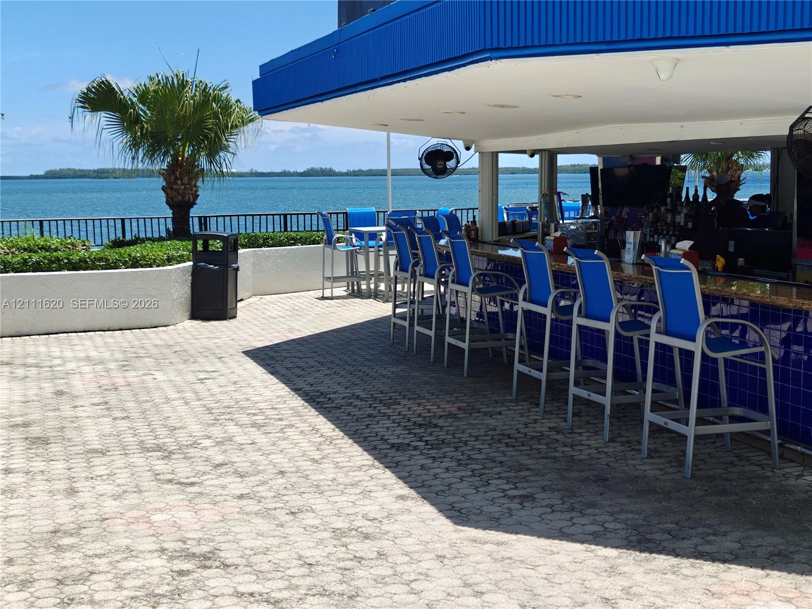 Outdoor bar featuring a covered serving area, blue tile facade, paver patio, and metal railing overlooking the water