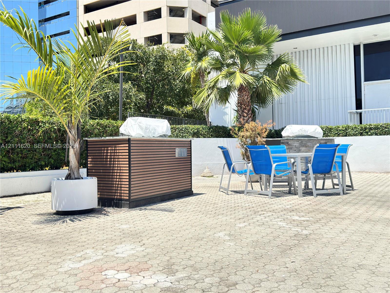 Outdoor terrace featuring stone paver flooring, built-in cabinetry with horizontal slat detailing, stone-accented grilling station, and natural privacy hedging