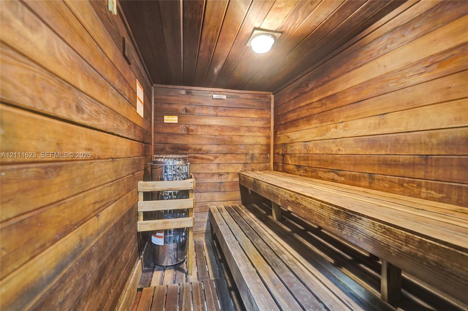 Sauna featuring horizontal wood-paneled walls and ceiling, multi-level slatted wood benches, and a metallic heater unit