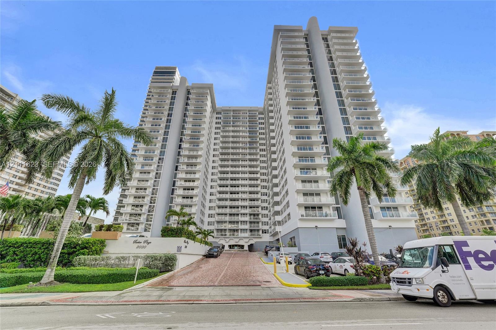 High-rise residential building featuring expansive private balconies, a paved drive-up entrance, and exterior tropical landscaping