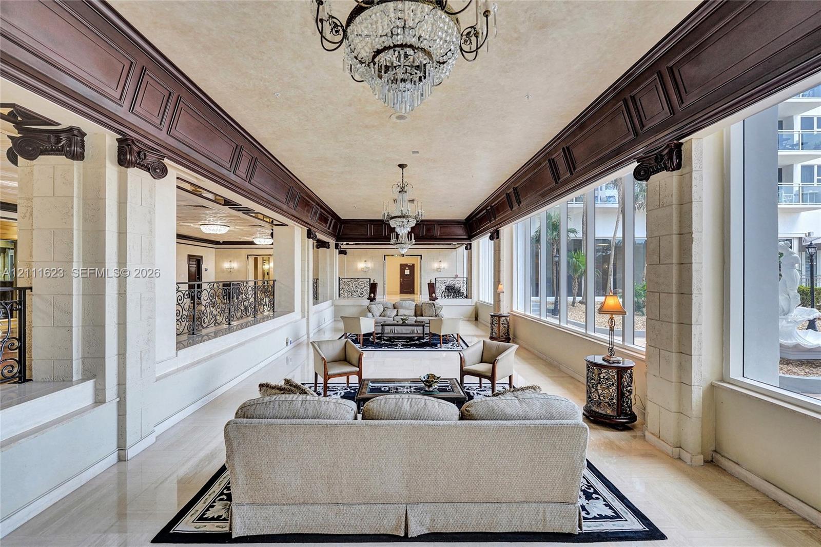 Expansive hall featuring ornate dark wood crown molding, stone-textured pillars, floor-to-ceiling windows, polished stone flooring, and tiered crystal chandeliers