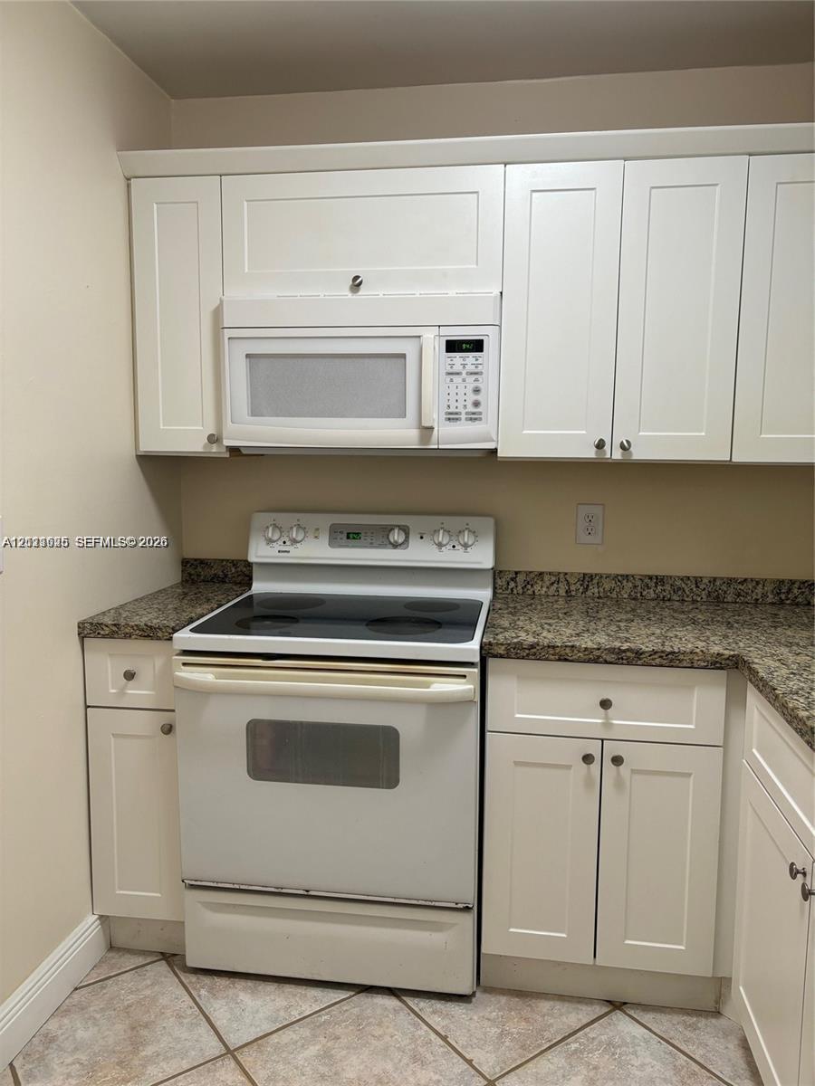 Kitchen featuring white shaker-style cabinetry, a stone-finish countertop, a built-in over-the-range microwave, a white electric range, and tile flooring