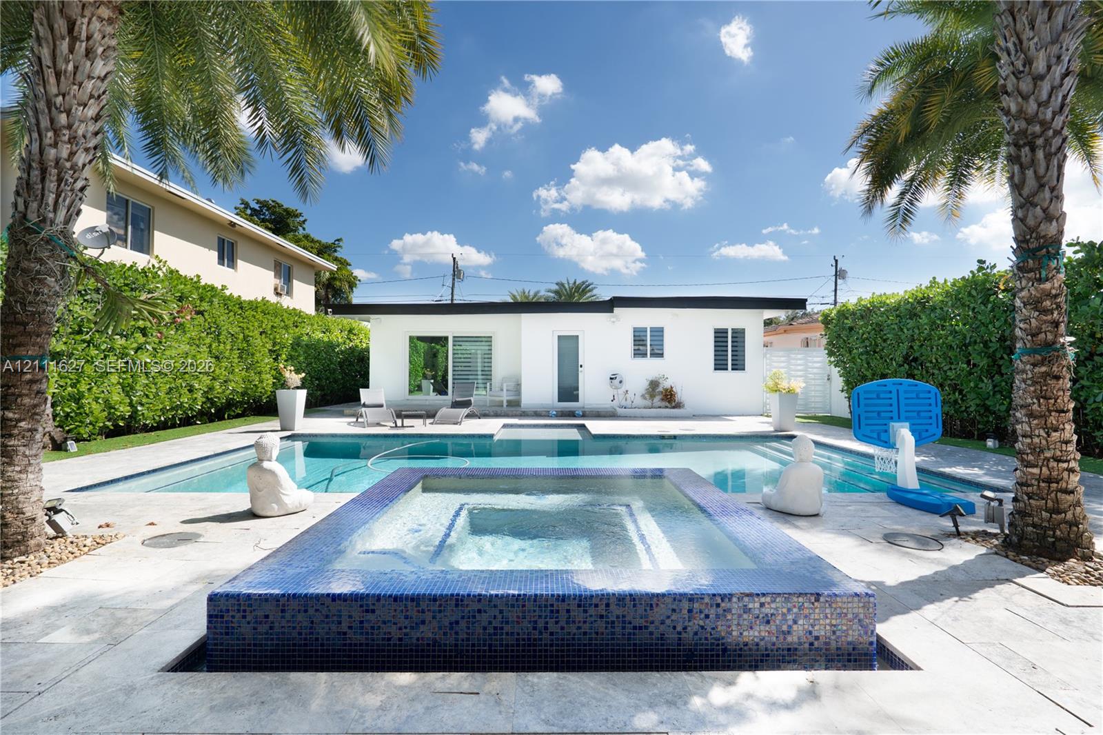 Exterior backyard space featuring an in-ground pool, a raised spa finished with blue glass mosaic tiles, light-toned stone patio surfacing, and privacy hedges