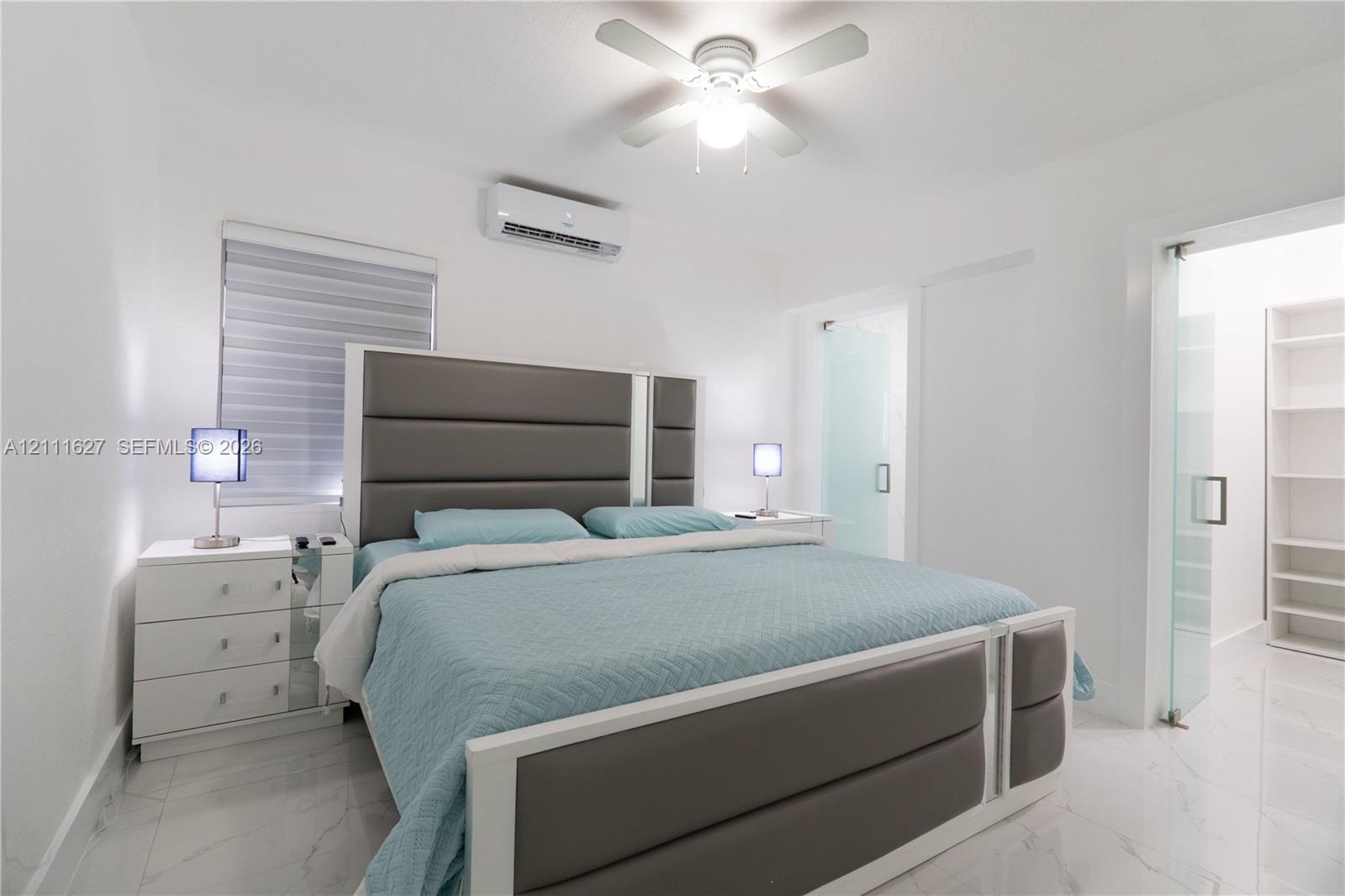 Neutral-toned interior featuring large-format tile flooring, a wall-mounted ductless air conditioning unit, a ceiling fan with integrated lighting, and a glass-paneled entry to a built-in shelving unit