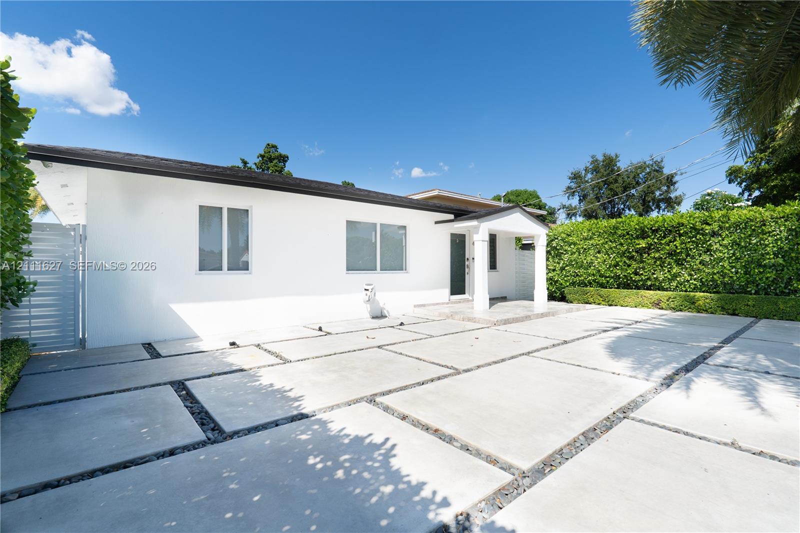 Single-story exterior featuring white stucco finish, large-format concrete paver driveway with decorative stone borders, covered entryway with support columns, and privacy hedging