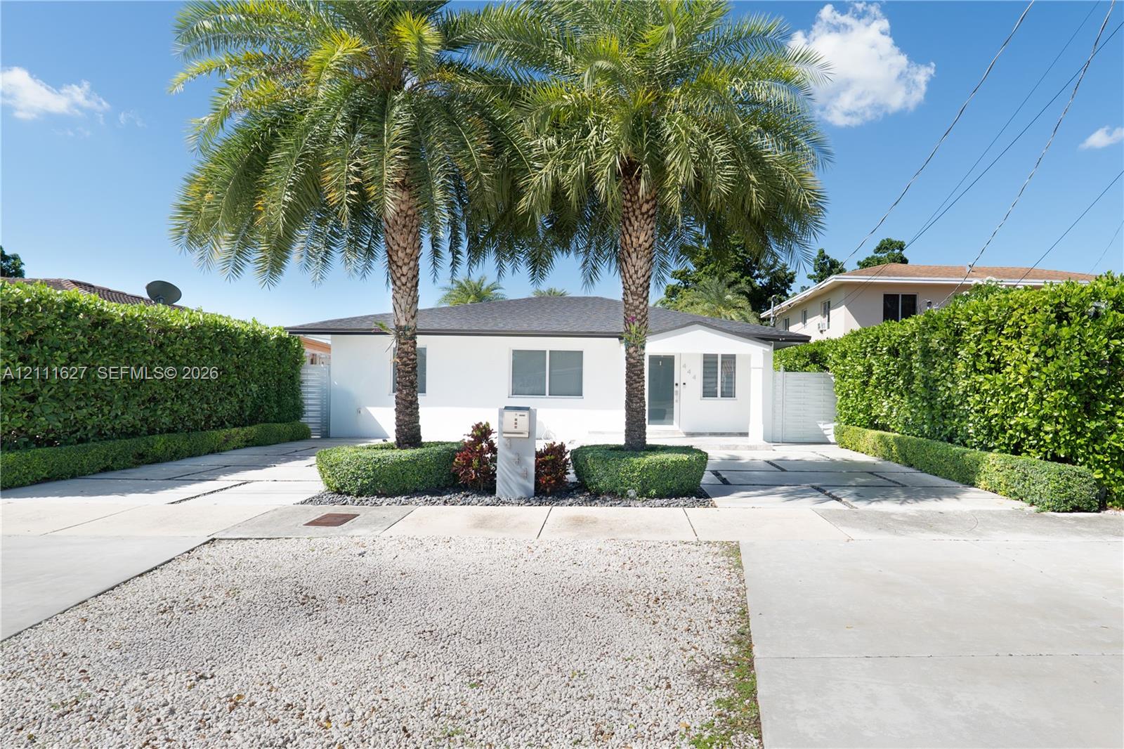 Single-story exterior featuring white stucco siding, a hip roof, expansive concrete paved driveway with integrated stone borders, mature palm trees, and dense perimeter hedge landscaping
