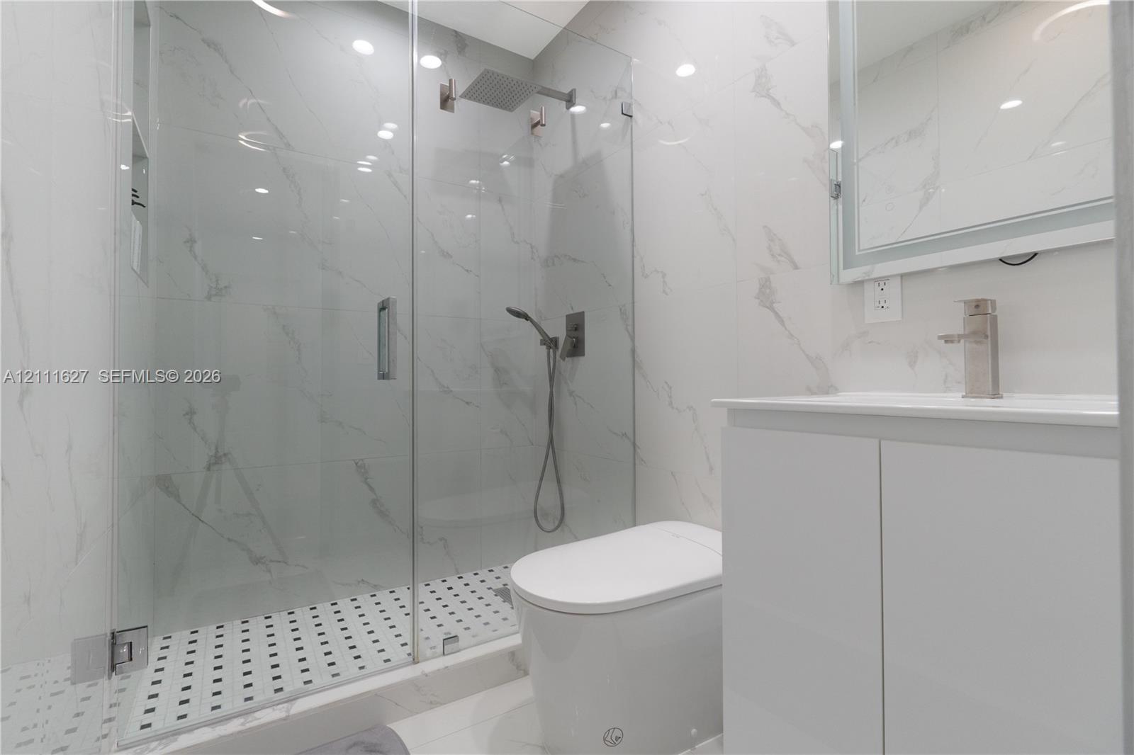 Walk-in glass-enclosed shower with a rainfall showerhead, large-format tile surround, mosaic tile flooring, and integrated wall niche, complemented by a white vanity with a metallic faucet and integrated lighting