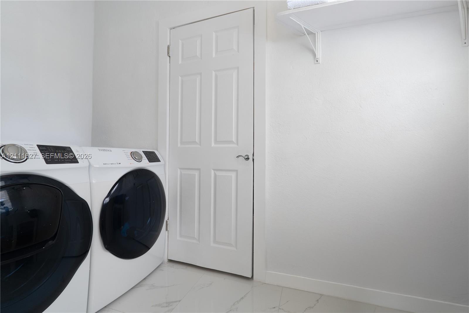 Laundry room featuring large-format tile flooring, a six-panel door with brushed nickel hardware, and an overhead built-in white storage shelf
