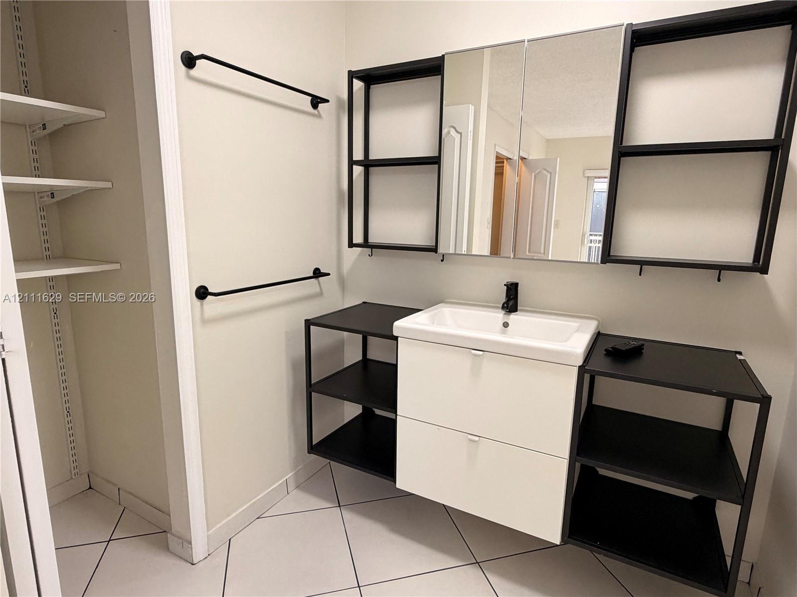 Wall-mounted vanity featuring a rectangular integrated sink, matte black faucet, open metal shelving, and white cabinetry, complemented by a tri-panel mirrored medicine cabinet and tiled flooring