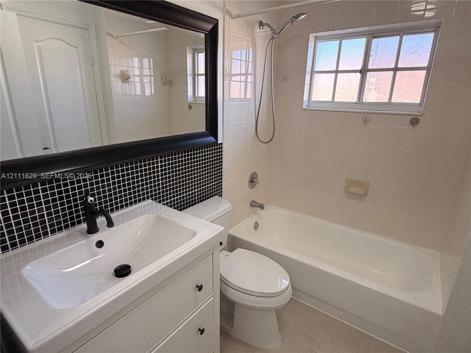 Bathroom featuring a white vanity with a rectangular sink, black faucet hardware, black-and-white grid tile backsplash, framed wall mirror, and a tub-shower combination with a sliding window