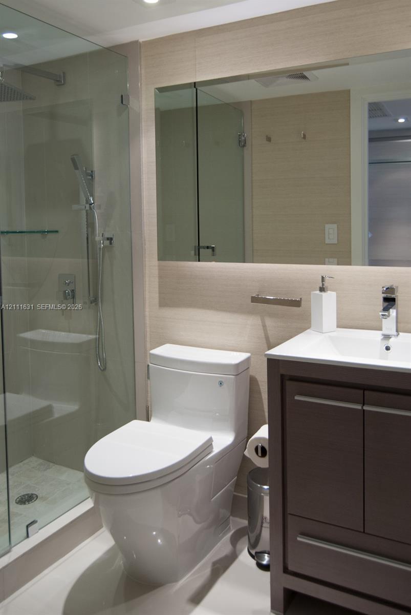 Bathroom featuring a glass-enclosed shower, wood-finish vanity with a rectangular sink, wall-mounted mirror, and neutral wall tile