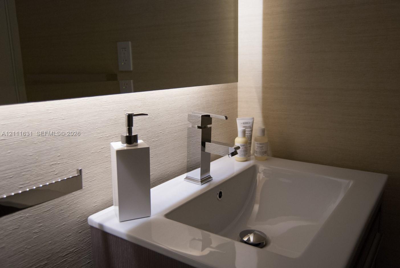 Undermount sink with integrated countertop, polished chrome single-lever faucet, wall-mounted mirror with integrated under-cabinet lighting, and textured wall treatment