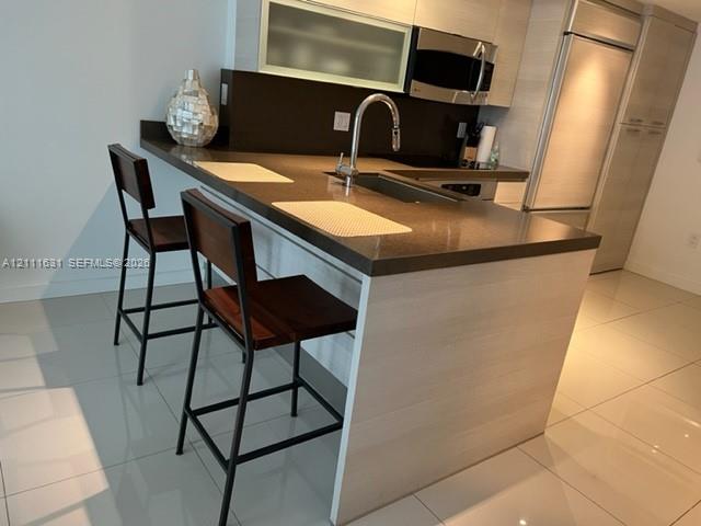 Kitchen featuring a peninsula with solid-surface countertops, horizontal-grain cabinetry, integrated refrigerator, stainless steel microwave, and polished tile flooring